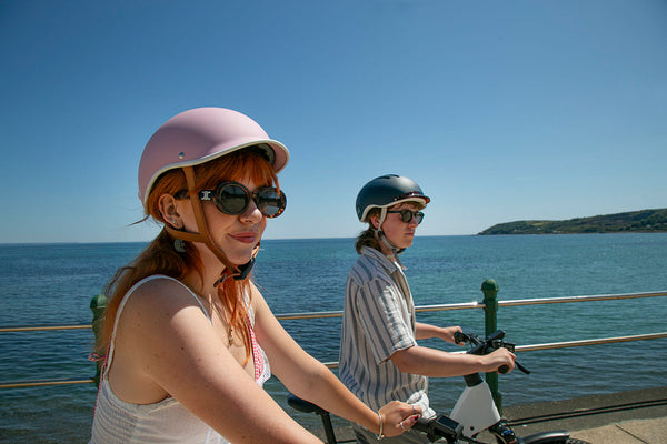 Two people riding electric bikes by a waterfront on a clear day.